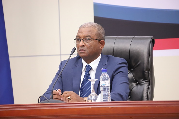 Homem de fato azul e gravata listrada sentado numa cadeira de couro preta, falando ao microfone durante uma conferência. À sua frente há uma garrafa de água e um copo.