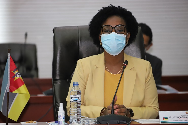 Mulher sentada à mesa de conferência, vestida com casaco amarelo e máscara cirúrgica, falando ao microfone. À sua frente há uma garrafa de água, um frasco de desinfetante e uma pequena bandeira de Moçambique.