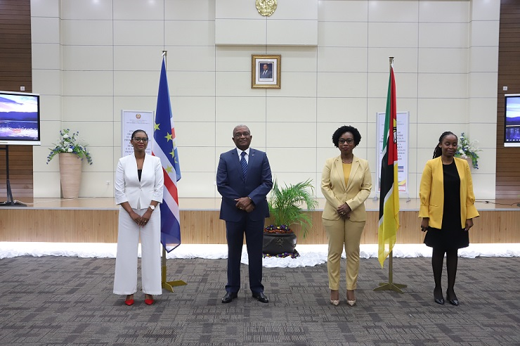 Quatro pessoas em pé, posando formalmente para uma fotografia num auditório. Ao centro, um homem de fato e gravata, ladeado por duas mulheres à esquerda e uma à direita. Atrás deles, as bandeiras de Cabo Verde e de Moçambique e uma parede clara com um retrato ao centro.